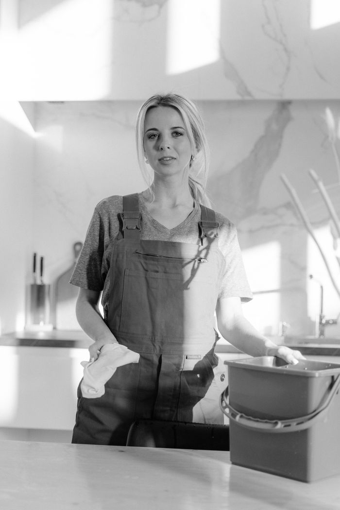about-us-01 Portrait en noir et blanc d’une femme de ménage avec des produits de nettoyage dans une cuisine moderne.